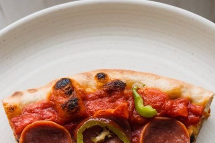 A pizza slice with an intricate tapestry-like pattern, topped with pepperoni and green peppers, on a white plate.