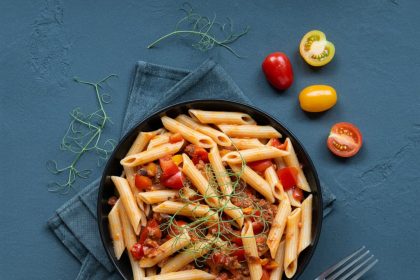 Overhead shot of a black bowl filled with penne pasta and meat sauce with colorful garnishes for a food photography prompt